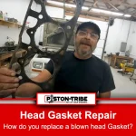 A bearded mechanic wearing a cap and glasses holds up a used head gasket in his right hand inside a workshop. The mechanic is standing near a white vehicle with its hood open, and various tools and equipment are visible in the background. The mechanic wears a navy blue t-shirt with the word 'Garage' printed on it, and the environment is brightly lit with a clean, organised workshop setting