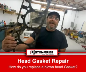 A bearded mechanic wearing a cap and glasses holds up a used head gasket in his right hand inside a workshop. The mechanic is standing near a white vehicle with its hood open, and various tools and equipment are visible in the background. The mechanic wears a navy blue t-shirt with the word 'Garage' printed on it, and the environment is brightly lit with a clean, organised workshop setting