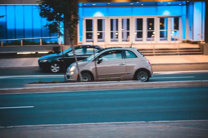 A Fiat 500 driving past a modern blue-lit building on a city street.