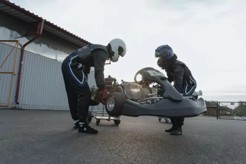 Two drivers in racing suits and helmets carry a go-kart for a race.