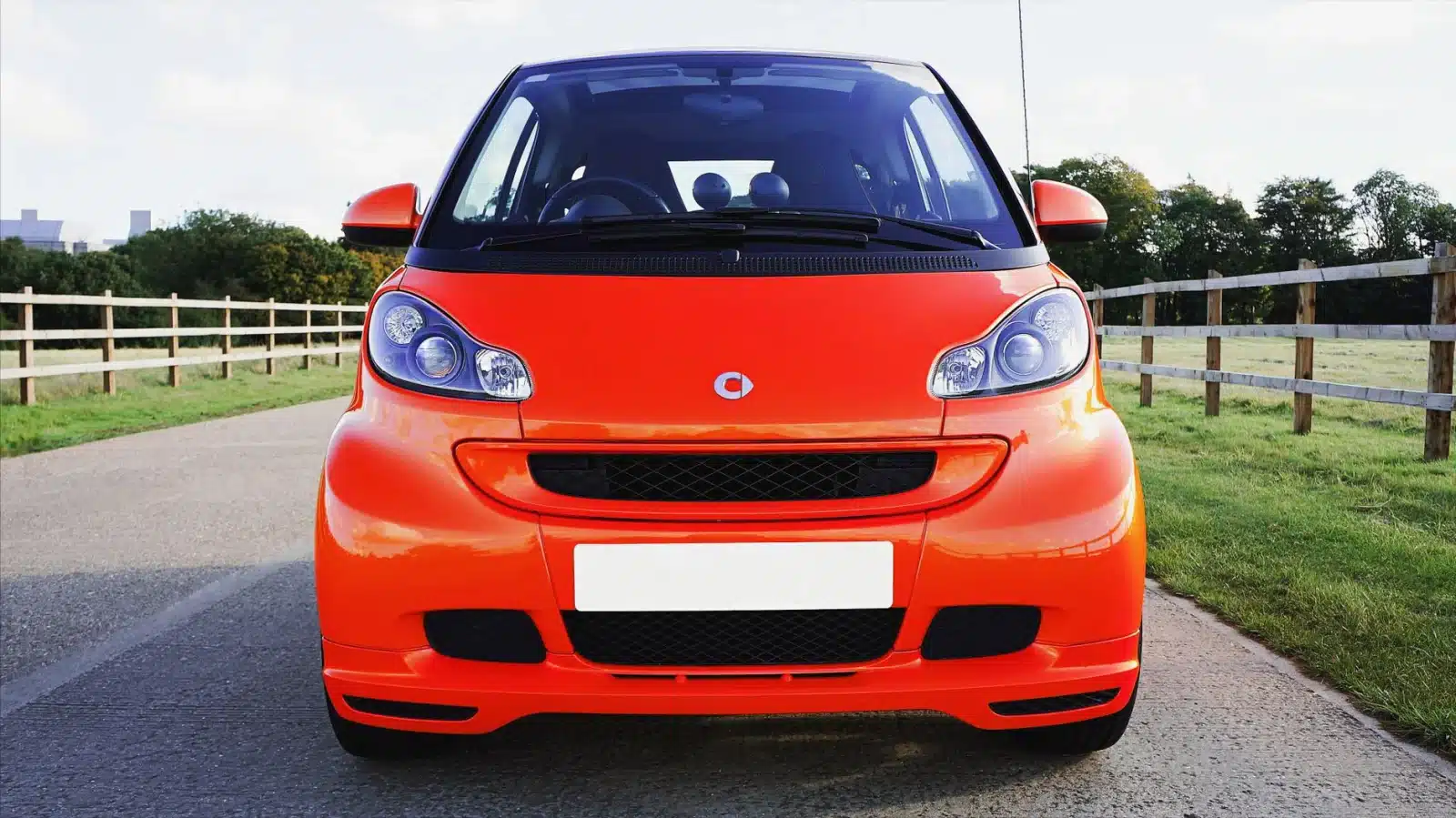Vibrant orange Smart car parked on a road in Knebworth, England, with wooden fences and green grass.