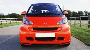 Vibrant orange Smart car parked on a road in Knebworth, England, with wooden fences and green grass.
