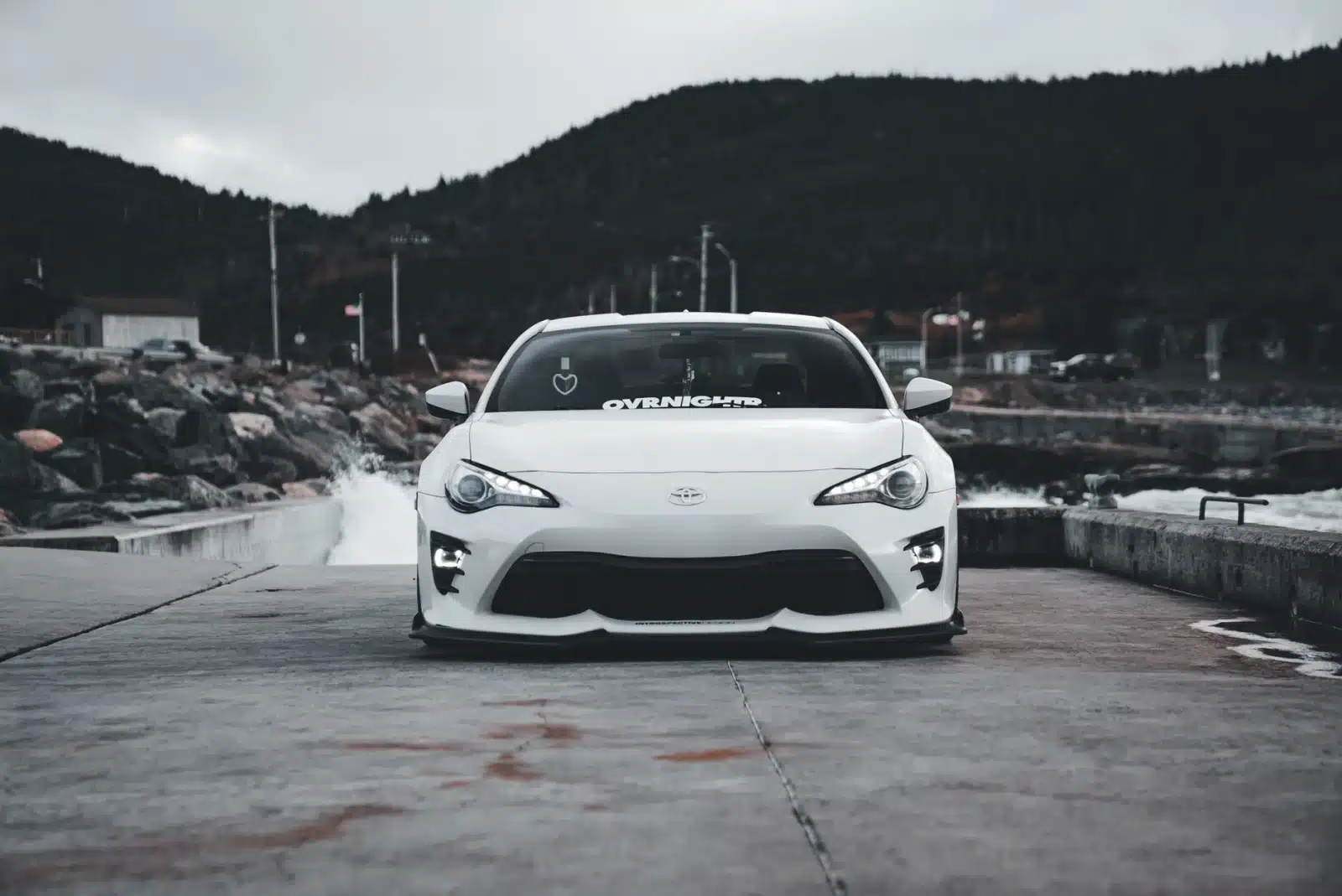 White sports car parked on a coastal road with an overcast sky background, exuding a cool and dramatic vibe.