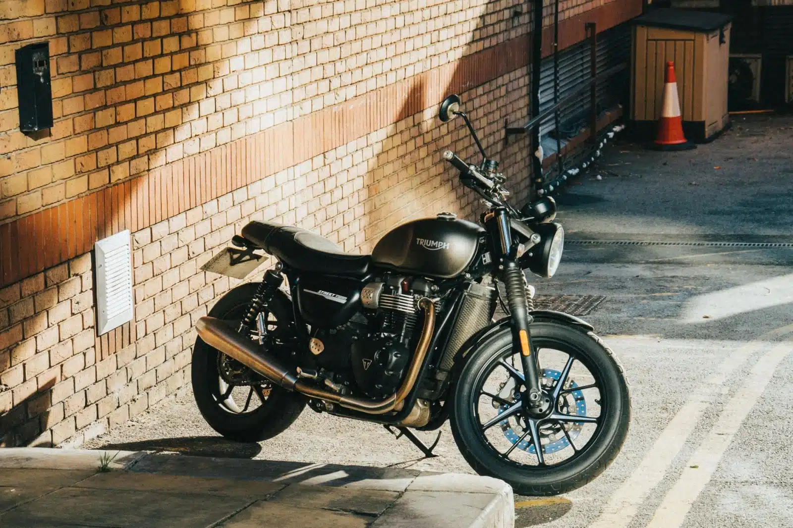 A classic Triumph motorcycle parked under sunlight in an urban alleyway with a brick wall background.