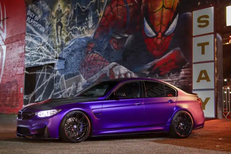 A striking purple BMW parked in front of a vibrant Spiderman mural at night, showcasing urban art and automotive design.