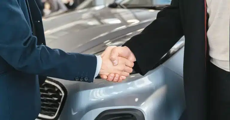 A close-up of two people shaking hands in front of a car in an indoor setting.
