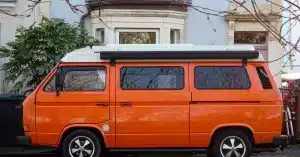 Side view of a classic orange van parked outdoors near a tree and house.